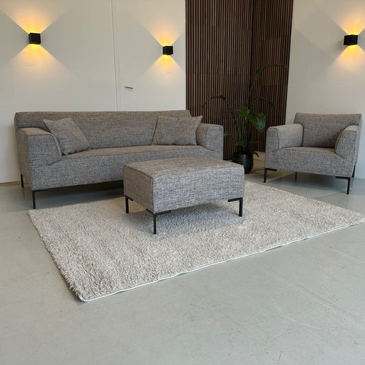 Gemêleerde Design on Stock Bloq zithoek met rechte bank, fauteuil en hocker, strakke lijnen en stevige kussens, opgesteld in een lichte showroom.