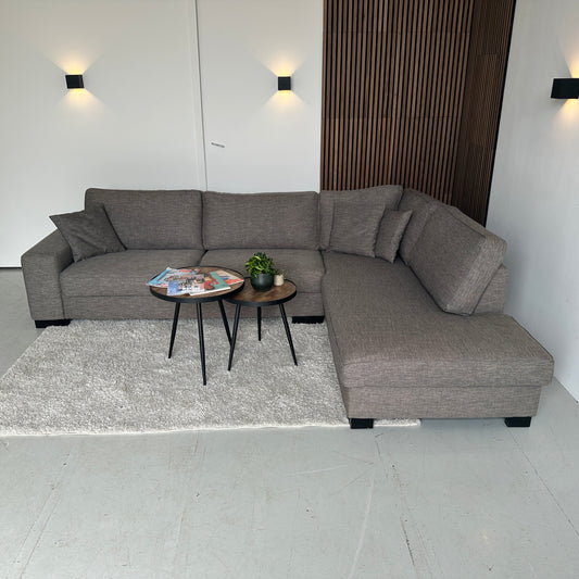 Gemêleerde taupe hoekbank van Goossens met zachte zit en losse kussens, opgesteld in een lichte showroom en klaar voor een tweede leven.