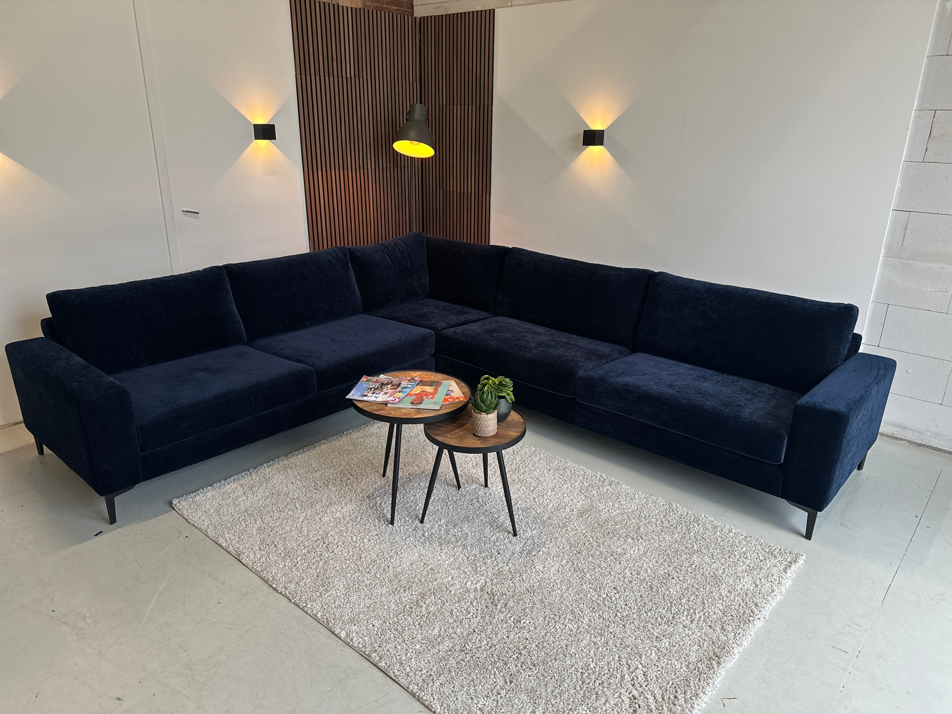 Donkerblauwe velours hoekbank Noah XL van INNO met royale opstelling, diepe zit en slanke poten, opgesteld in een lichte showroom.